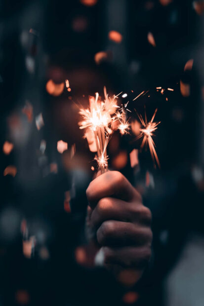 A New Year Aesthetic Wallpaper featuring a hand holding a glowing sparkler surrounded by blurry confetti and dark background