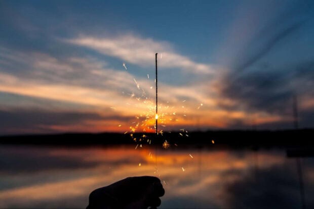 A beautiful New Year aesthetic wallpaper showing a hand holding a bright sparkler against a vibrant sunset sky reflecting over a calm lake