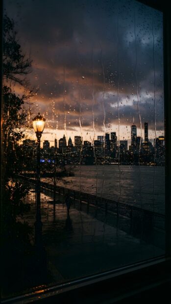A captivating rain wallpaper showing a city skyline across the water, viewed through a window with rain streaks. A glowing street lamp illuminates the foreground during dusk