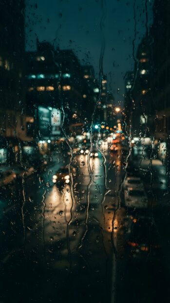 A moody Rain Wallpaper capturing a night city street scene through a window covered in raindrops, creating blurred lights and a dark, atmospheric view. The bokeh effect of car headlights and streetlights adds depth