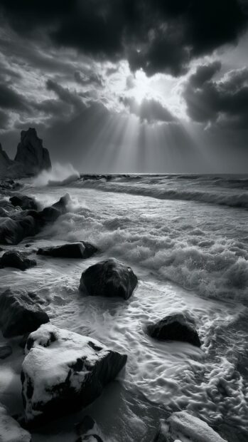 A dramatic black and white winter wallpaper showing a stormy ocean with sun rays breaking through dark clouds over snow-covered rocks
