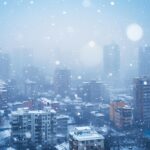 Cloudy winter cityscape with snow falling over high-rise buildings and residential apartments.