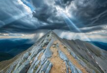 Majestic mountain summit with rocky trail under dark storm clouds and rays of sunlight breaking through, showcasing a dramatic and awe-inspiring landscape.