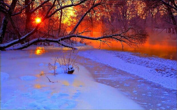 Snow-covered riverbank with leafless trees and a vibrant sunset casting orange and purple hues over the frozen water.
