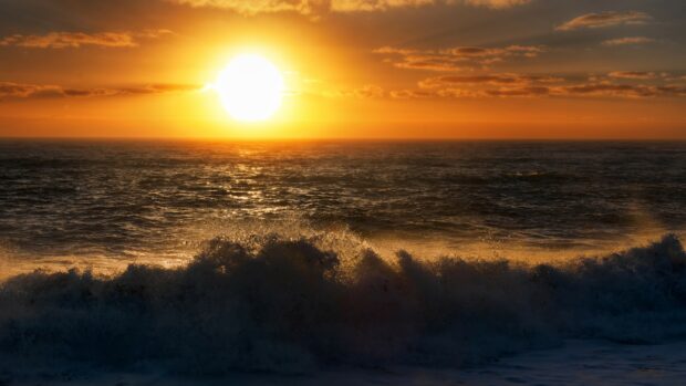 Vivid sunset over the ocean with waves crashing on shore and a mostly cloudy sky.