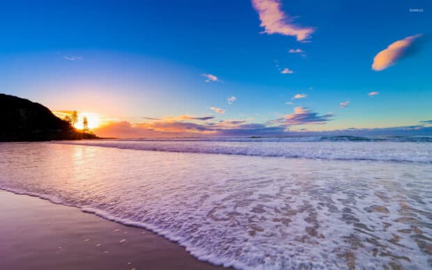 Vibrant sunset over a calm beach with gentle waves and a tree-lined hill in the background.