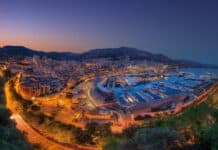 Luxury cityscape of Monte Carlo marina at dusk with yachts, illuminated buildings, and surrounding hills on a clear evening.