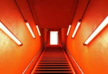 Vivid orange staircase with metal handrails leading to a bright white door, illuminated by four fluorescent lights on textured orange walls.